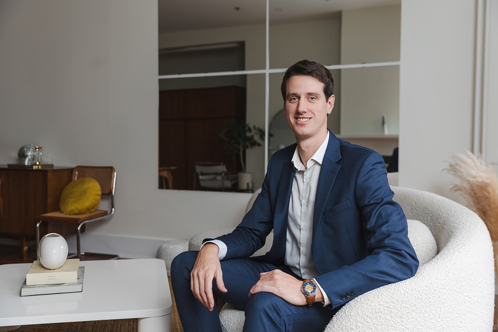 A man in a blue suit sits in a modern, white armchair in a stylish living room, smiling at the camera—perhaps discussing whole life insurance or policy loans. A coffee table with books and a decorative item sits in the foreground.