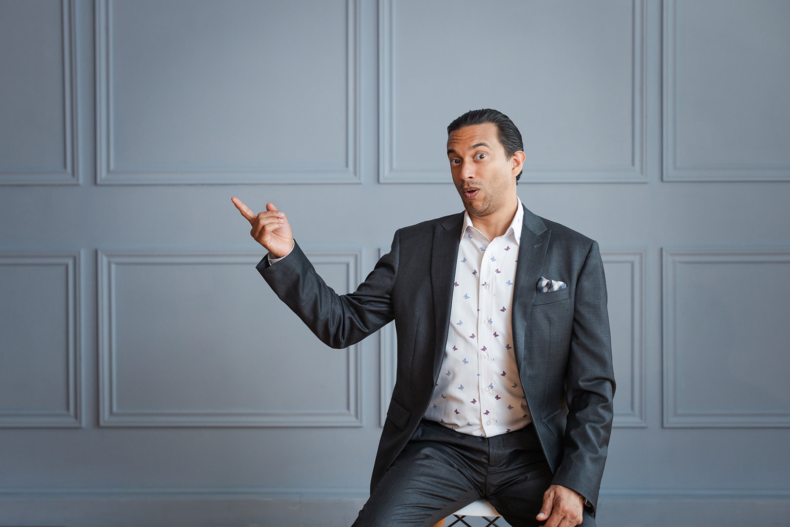 A man in a dark suit sits on a stool against a gray paneled wall, gesturing animatedly to his left with his index finger while making a surprised facial expression.